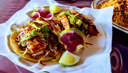 Two tacos topped with grilled chicken, avocado, and cabbage, served with lime wedges and colorful radish slices.