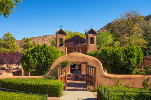 A scenic view of a historic adobe church surrounded by lush greenery and mountains under a clear blue sky.