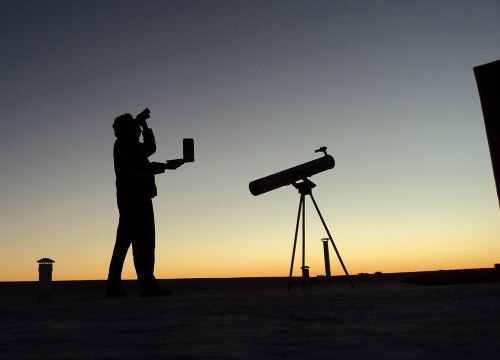 A silhouette of a person holding a camera near a telescope against a sunset sky.