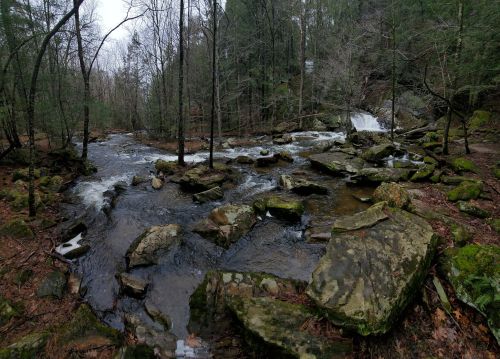 A serene river flows through a rocky landscape, surrounded by trees and a gentle waterfall in the background.