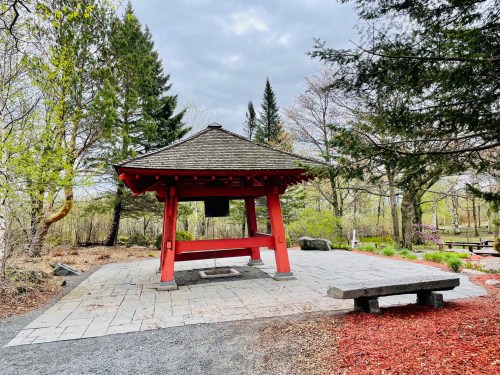 A red gazebo with a sloped roof stands on a stone patio, surrounded by trees and landscaped greenery.