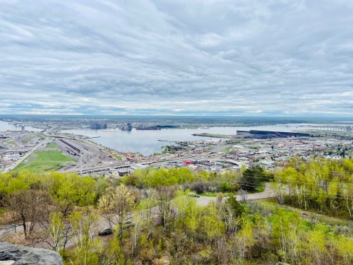 A panoramic view of a river and industrial area, surrounded by greenery under a cloudy sky.