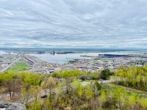 A panoramic view of a river and industrial area, surrounded by greenery under a cloudy sky.