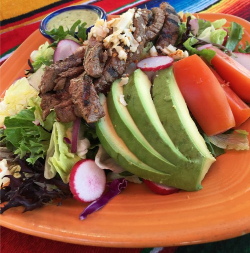 A vibrant salad topped with grilled steak, avocado, tomatoes, radishes, and cheese, served on an orange plate.