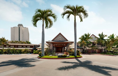 A tropical hotel entrance with palm trees and a modern building in the background under a clear blue sky.
