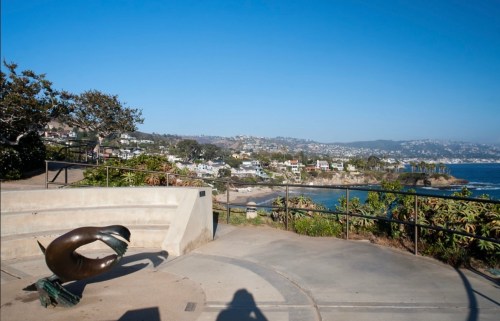 A coastal view featuring a sculpture, lush greenery, and homes along the shoreline under a clear blue sky.