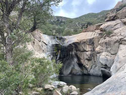 A serene waterfall cascades over rocky cliffs into a calm pool, surrounded by lush greenery and hills in the background.