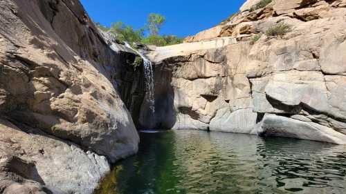 A serene waterfall cascading into a clear pool, surrounded by rocky cliffs and greenery under a bright blue sky.