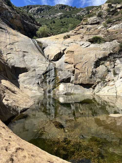 A serene rocky landscape with a small waterfall cascading into a clear pool, surrounded by greenery and blue sky.