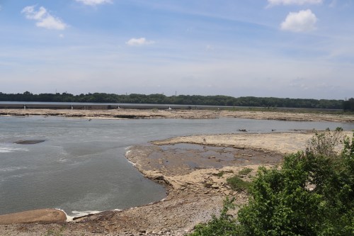 A wide river with rocky banks under a blue sky, surrounded by greenery and distant trees.