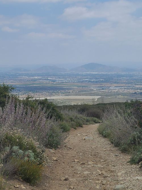 A dirt path leads through greenery, overlooking a valley with distant mountains under a cloudy sky.