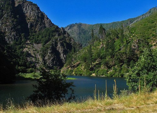 A serene lake surrounded by lush green mountains and trees under a clear blue sky.