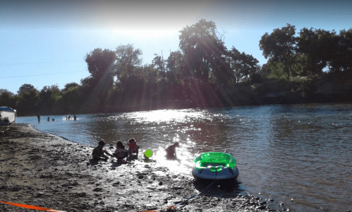 A sunny riverside scene with people playing in the water and a green inflatable raft on the shore.