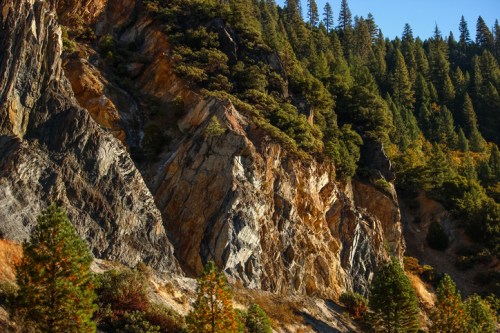 Rocky cliffside with lush green trees and autumn foliage under a clear blue sky.