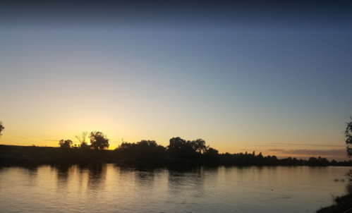 A serene river at sunset, with silhouettes of trees and a colorful sky reflecting on the water's surface.