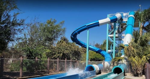 A colorful water slide spirals down into a splash pool, surrounded by greenery and a clear blue sky.