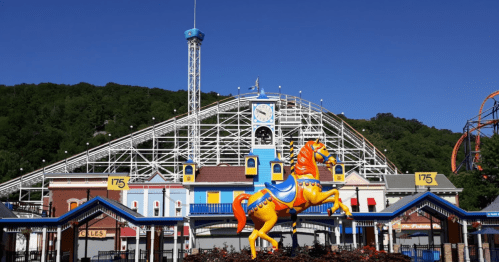 Colorful carousel horse in front of a roller coaster and amusement park buildings under a clear blue sky.