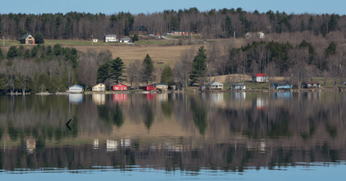 A serene lake scene with colorful houses reflecting on the water, surrounded by trees and rolling hills.