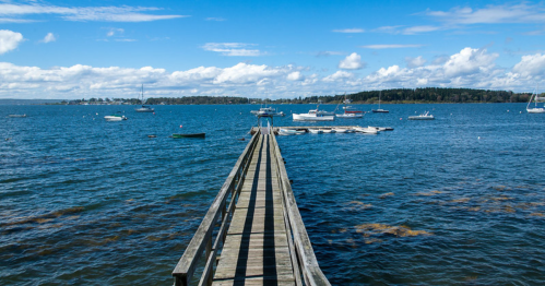 A wooden pier extends into a calm blue sea, with boats floating and clouds scattered in the sky.