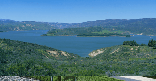 A panoramic view of a serene lake surrounded by green hills and mountains under a clear blue sky.