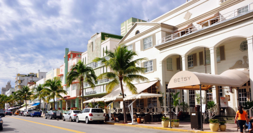 Colorful buildings line a palm tree-lined street, with outdoor seating and a sign for the Betsy hotel.