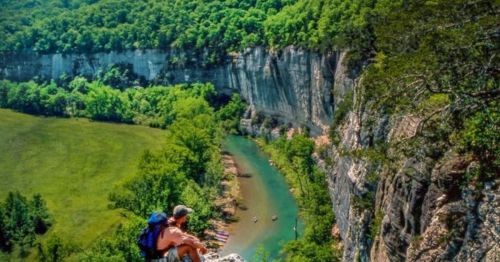 A hiker sits on a cliff overlooking a lush green valley and a winding river below.