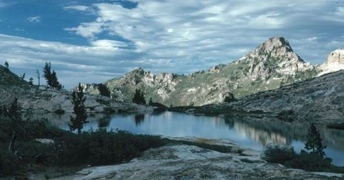 A serene mountain landscape with a calm lake reflecting the sky and rugged peaks under a cloudy blue sky.
