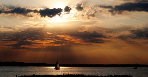 A serene sunset over a calm lake, with a sailboat silhouetted against golden and orange clouds.