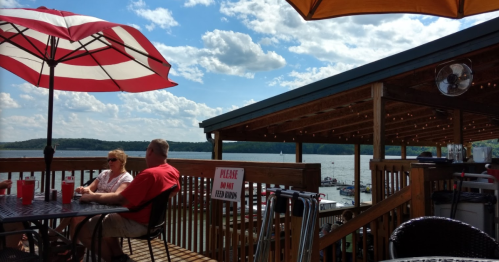 A couple sits at a patio table under a red and white umbrella, overlooking a lake with boats and a cloudy sky.