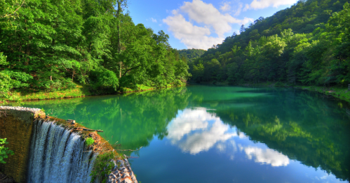 A serene landscape featuring a calm, reflective lake surrounded by lush green trees and a small waterfall.