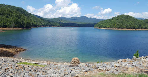 A serene lake surrounded by green hills and mountains under a blue sky with scattered clouds.