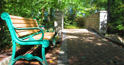A wooden bench with green metal arms beside a stone bridge over a brick path in a lush, green park.