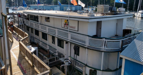 A large houseboat docked at a marina, with flags flying and a wooden walkway leading to it.