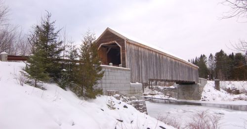 A wooden covered bridge spans a frozen river, surrounded by snow-covered trees and a winter landscape.