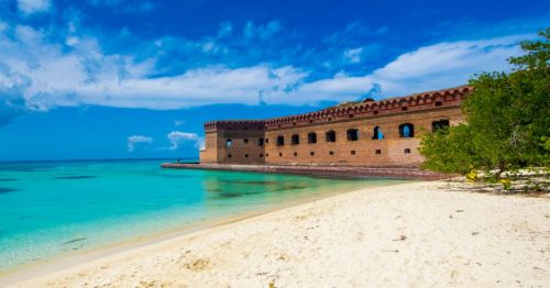 A historic brick fort by a sandy beach, surrounded by clear turquoise waters and a blue sky with scattered clouds.