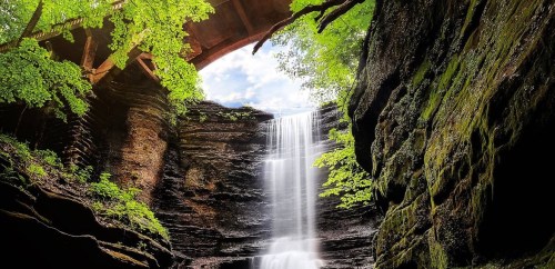 A serene waterfall cascades down rocky cliffs, surrounded by lush greenery and a wooden bridge overhead.