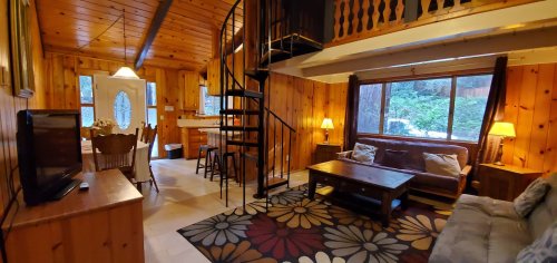 Cozy wooden cabin interior with a spiral staircase, living area, dining space, and large windows overlooking greenery.