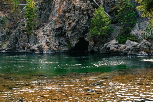 A serene river scene with a rocky cave entrance, surrounded by lush green trees and calm water reflecting the landscape.