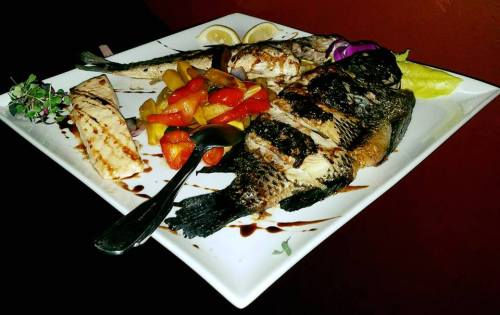 A plate of grilled fish garnished with lemon, colorful peppers, and herbs, served with a spoon on a white dish.