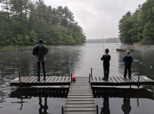 Two children fishing from wooden docks on a misty lake surrounded by trees.