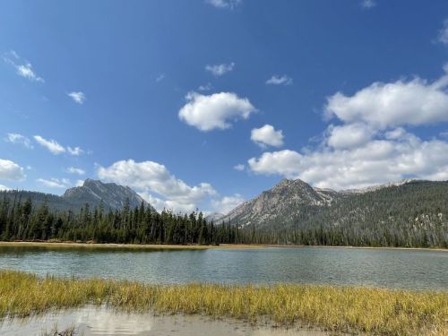 A serene landscape featuring a lake surrounded by mountains and lush green trees under a blue sky with fluffy clouds.
