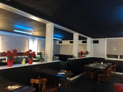 Interior of a restaurant featuring dark walls, wooden tables, and potted flowers, with soft lighting and window blinds.