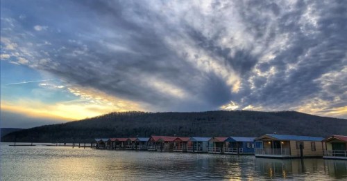 A serene lake scene with colorful cabins, dramatic clouds, and a sunset over a wooded hillside.