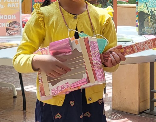 A child in a yellow cardigan holds a colorful, handmade guitar, smiling in a bright, sunny environment.