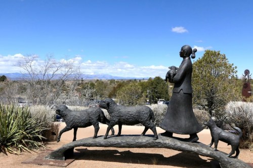 A bronze statue of a woman with sheep and a dog, set against a blue sky and distant mountains.