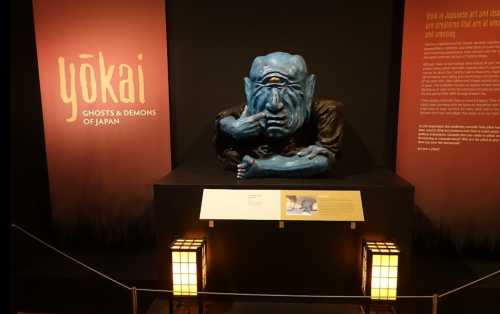 A blue, mythical creature sculpture with one eye, displayed in a museum about Japanese yōkai, with informative text panels.