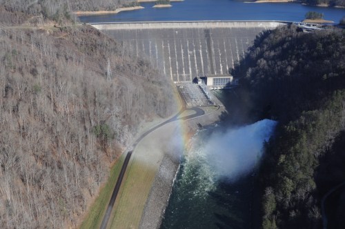 Aerial view of a dam with water flowing, surrounded by trees and a rainbow forming in the mist.