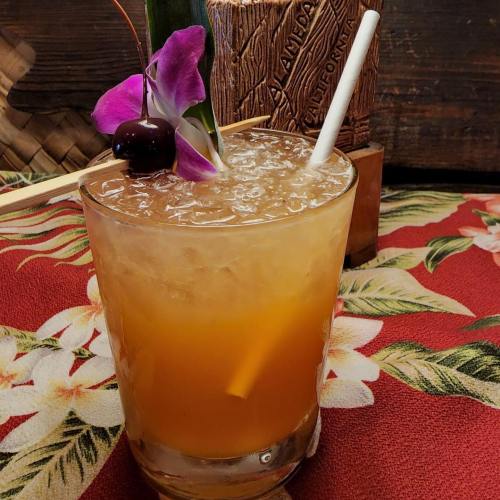 A tropical cocktail garnished with a purple flower and cherry, served in a glass on a floral tablecloth.