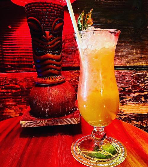 A tropical cocktail in a tall glass with ice, next to a carved tiki mug, set against a warm, rustic background.