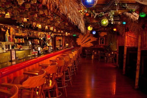 A vibrant tiki bar with a thatched roof, colorful lanterns, and a long wooden bar lined with stools.